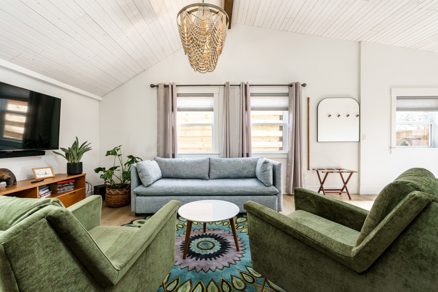 A cozy living room is highlighted by vaulted ceilings and natural light from the windows. A light blue sofa and two green chairs surround a circular coffee table, resting on a colorful area rug. Potted plants add a touch of greenery to the space.