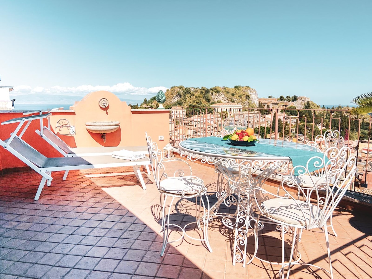 A spacious terrace features a round dining table surrounded by ornate chairs. A lounge chair is positioned nearby, all set against a sunny backdrop. A fruit bowl adds a pop of color to the table while offering views of the surrounding landscape.