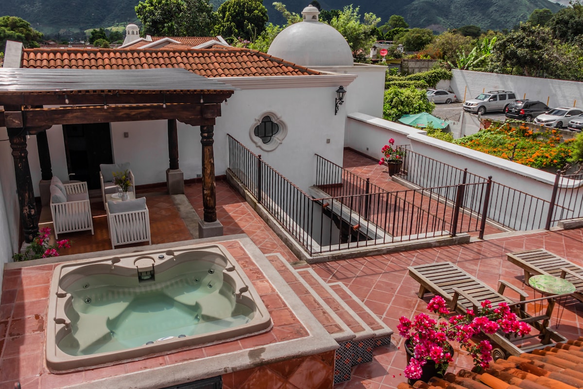 The spacious terrace features a relaxing jacuzzi surrounded by colorful flowers. Several lounge chairs are positioned beside the jacuzzi, offering a comfortable area to unwind. The views of the surrounding landscape and nearby mountains enhance the tranquil atmosphere of this outdoor space.