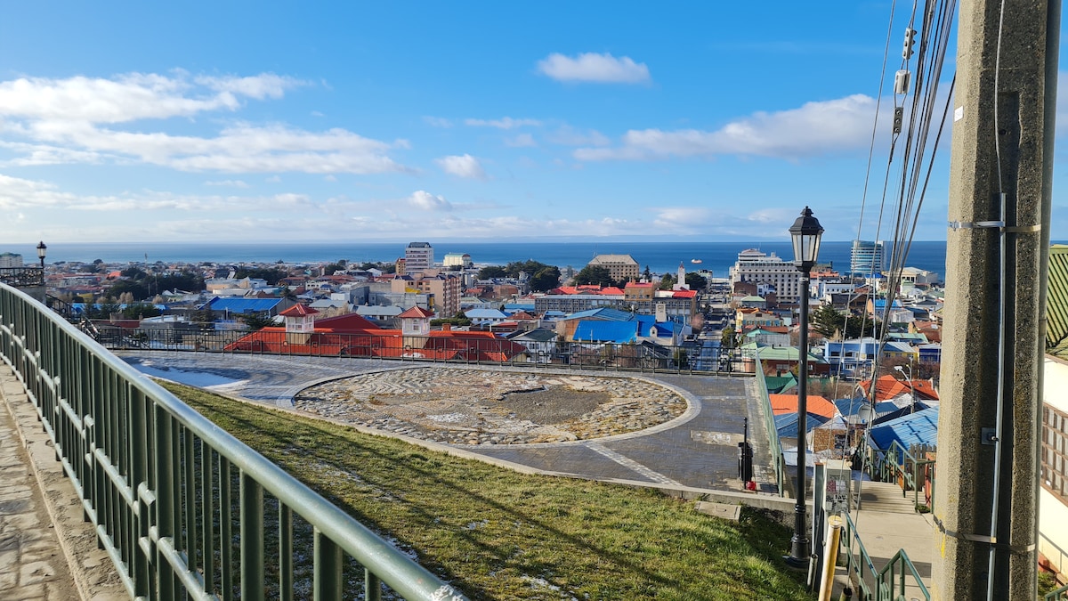 A scenic view captures a vibrant urban landscape with colorful rooftops and the ocean in the background. A circular stone plaza is seen in the foreground, framed by a railing, under a clear blue sky dotted with clouds.