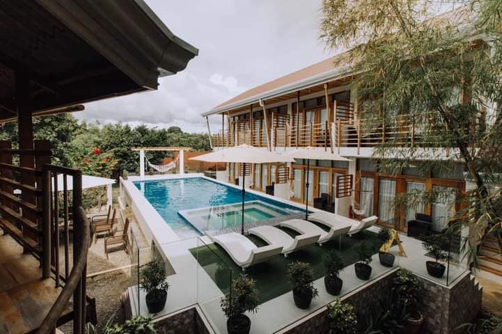 A modern tropical resort features an inviting pool surrounded by lounge chairs. The two-story building showcases airy balconies with wooden railings. Lush greenery and decorative plants are visible, enhancing the outdoor area, while a shaded seating space is positioned near the poolside.