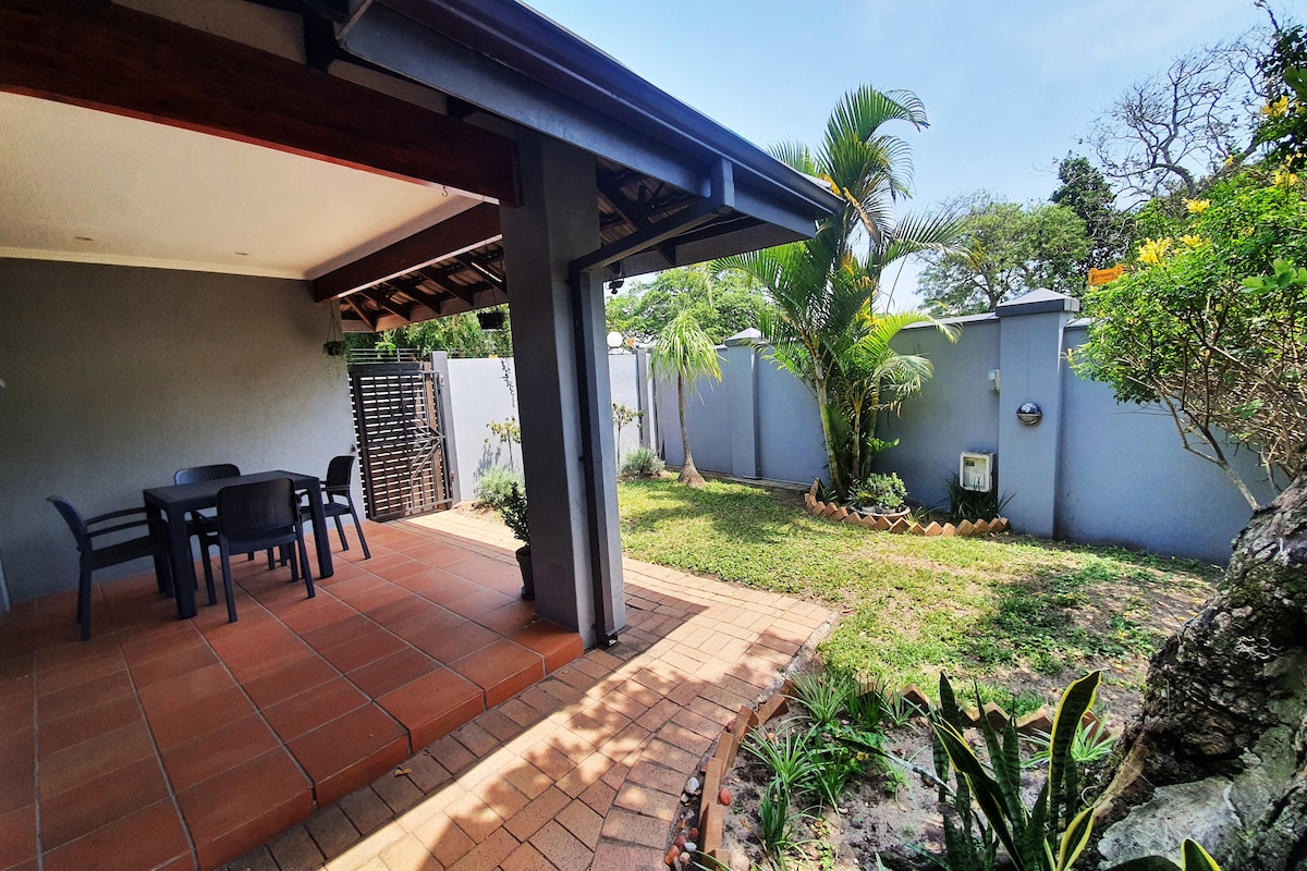An undercover patio area is visible, featuring a dining table with four chairs on a tiled surface. A small garden with grass and various plants is seen beyond the patio, bordered by a grey wall. The sky is clear, adding to the open feel of the space.