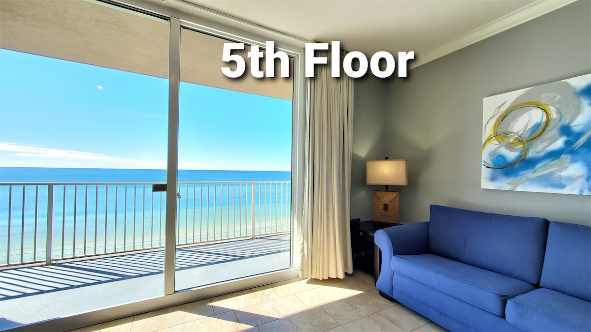 A bright living area showcases a blue sofa facing large floor-to-ceiling windows. The view reveals the expansive ocean beyond, with clear blue skies meeting the water. Soft curtains frame the windows, enhancing the view of the gulf from the fifth floor.