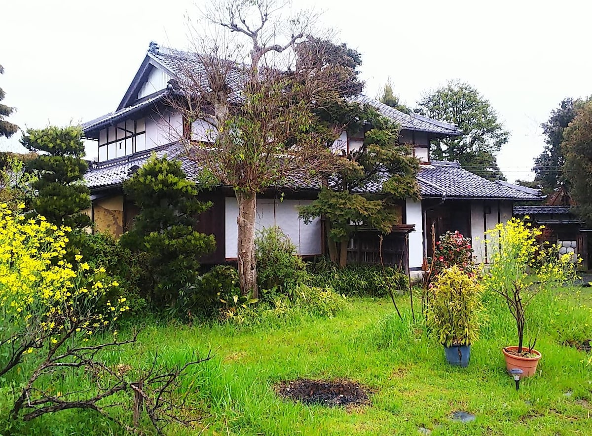 A traditional Japanese house is surrounded by a verdant garden, featuring various trees and flowering plants. The structure showcases a steeply pitched roof and sliding doors, emphasizing its classic architectural style. Lush greenery envelops the area, suggesting a peaceful retreat.