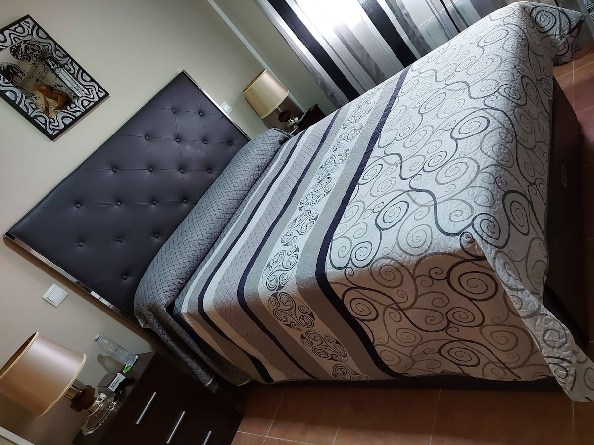A well-appointed bedroom features a spacious bed with an elaborately patterned duvet. Two bedside lamps provide soft illumination, and an abstract artwork adorns the wall above the headboard. Light filters through sheer drapes, creating a calm and inviting ambiance.