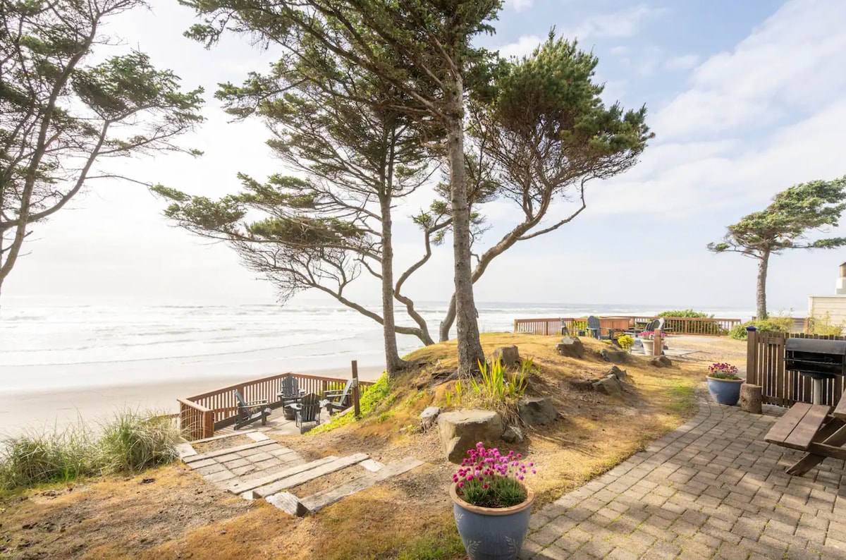 The outdoor area offers a scenic view of the beach, framed by tall trees swaying gently in the breeze. A stone pathway leads down towards the sandy shore, with comfortable seating arrangements visible on a wooden deck, surrounded by lush greenery and colorful planters.