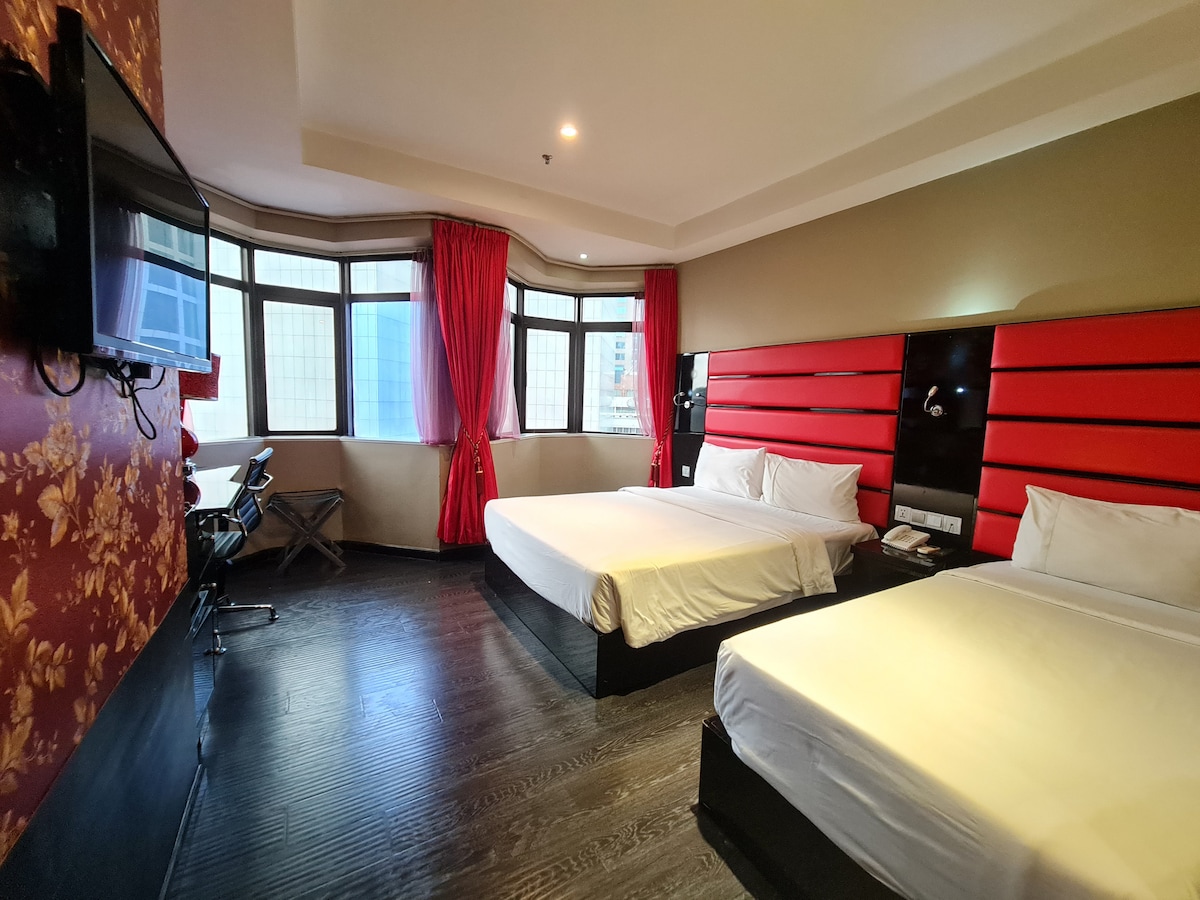 A well-appointed room features two spacious beds with a bold red headboard. Natural light floods the area through large windows, complemented by elegant red curtains. A functional workspace and a television can be seen on the wall, enhancing the room's practicality.