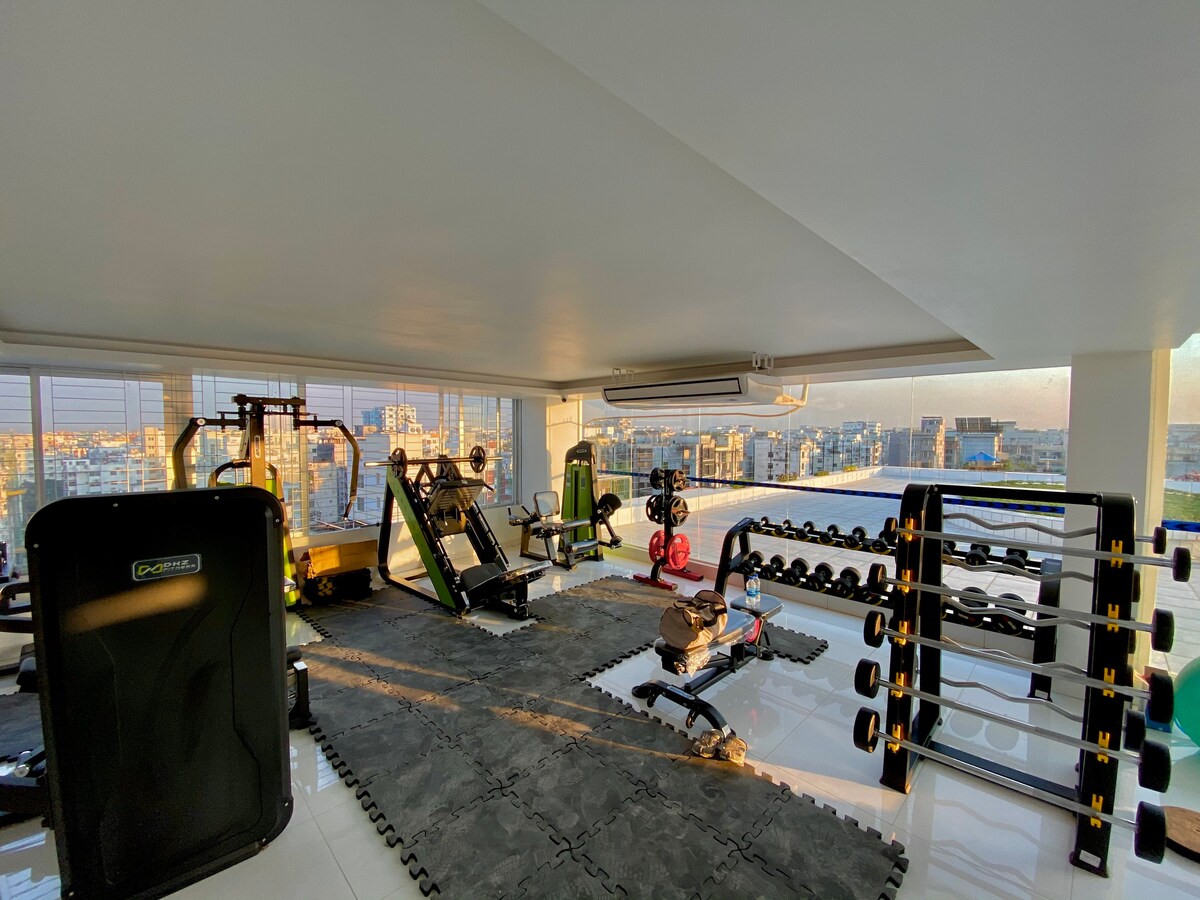 A well-equipped gym is presented with various fitness machines, free weights, and exercise mats arranged neatly on a glossy floor. Large windows allow ample natural light, offering views of the city skyline, enhancing the workout atmosphere.