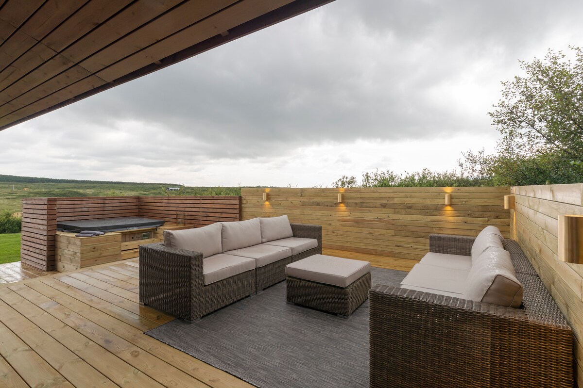 A spacious outdoor seating area features comfortable wicker sofas and a central coffee table, all arranged over wooden decking. Soft lighting illuminates the space, which is surrounded by natural greenery. A hot tub is positioned nearby, enhancing outdoor relaxation options.