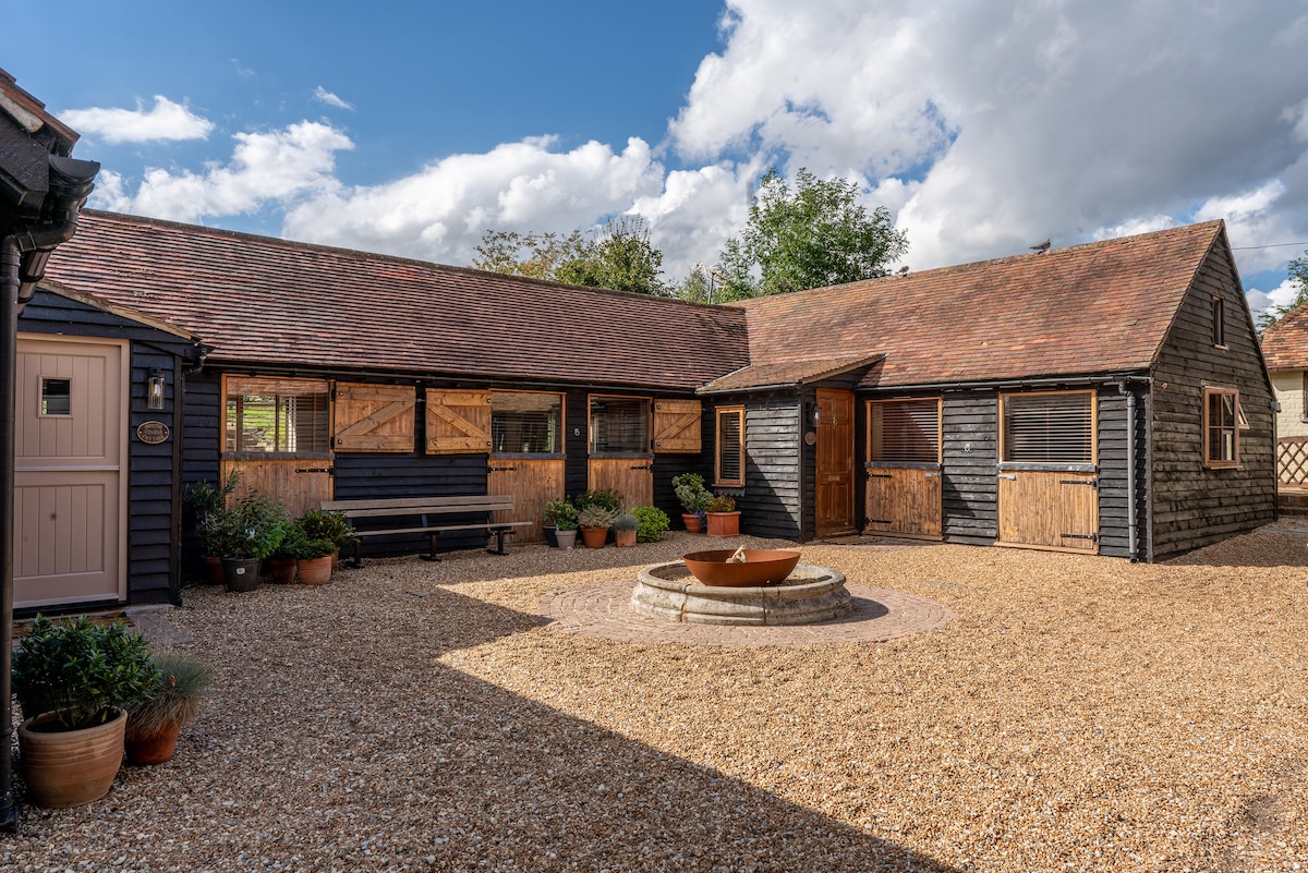 The exterior of a black wooden stable is showcased, featuring a gravel courtyard with potted plants. A circular stone feature is visible at the center, with benches placed along the walls. Large windows offer glimpses into the stable's interior.