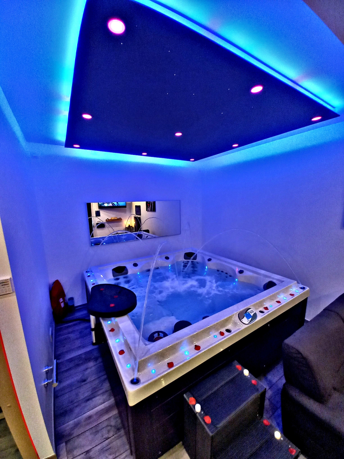 The image showcases a luxurious spa area featuring a spacious jacuzzi surrounded by ambient lighting and decorative accents. Water cascades gracefully from spouts, while a large mirror reflects the serene environment. A comfortable chair is positioned nearby for relaxation.