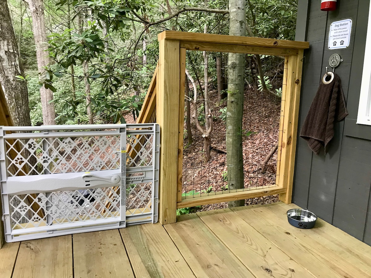 A wooden deck area features a wooden gate with a secure latch and a non-slip surface. A pet bowl is positioned on the floor, and a towel is hung on a hook nearby. Lush greenery is visible through the open gate, enhancing the natural setting.