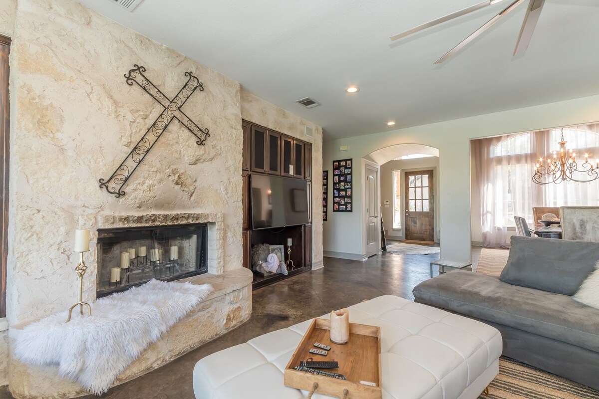 A stylish living area is highlighted by a stone wall with a decorative cross and a modern fireplace. A spacious sectional sofa faces a large television, while a plush white fur blanket rests on an oversized ottoman. Natural light flows through nearby windows, enhancing the relaxed setting.