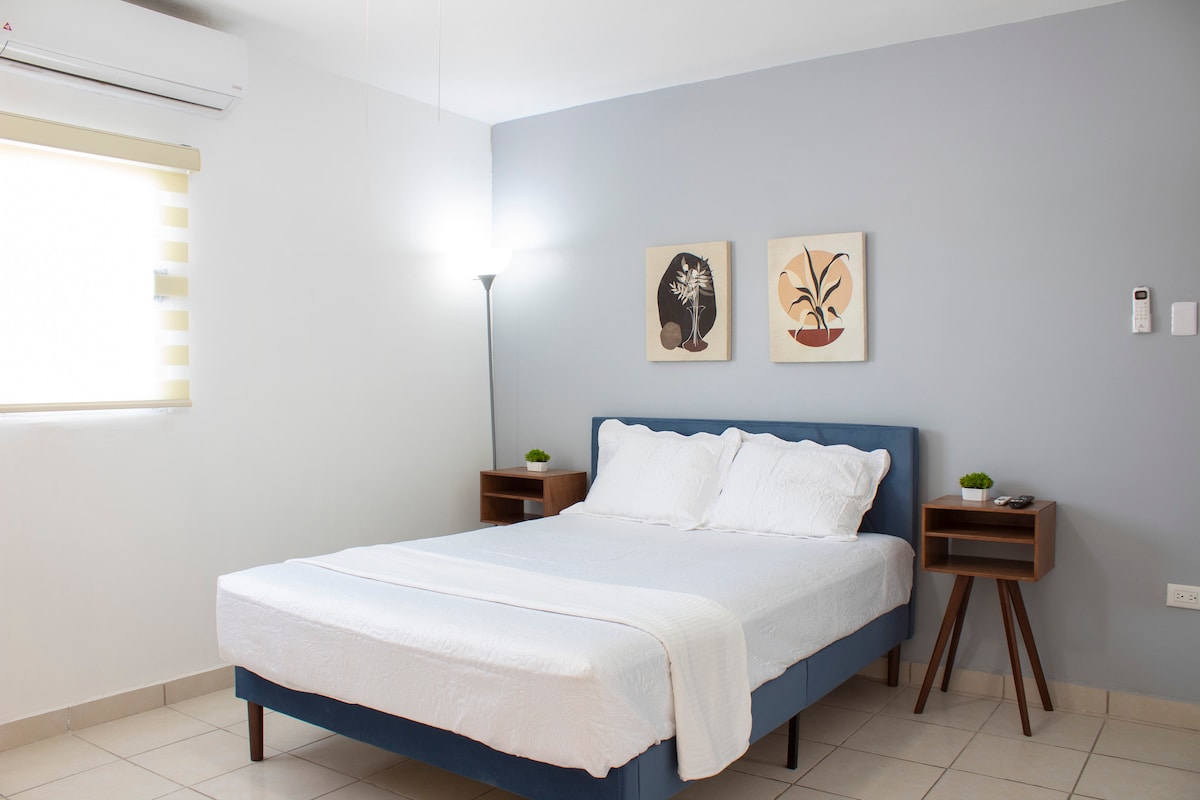 A modern bedroom features a spacious bed with white linens against a light grey wall. Two framed botanical artworks are positioned above the bed. Nightstands with small green plants are located on either side, and a window is adorned with light-filtering shades.