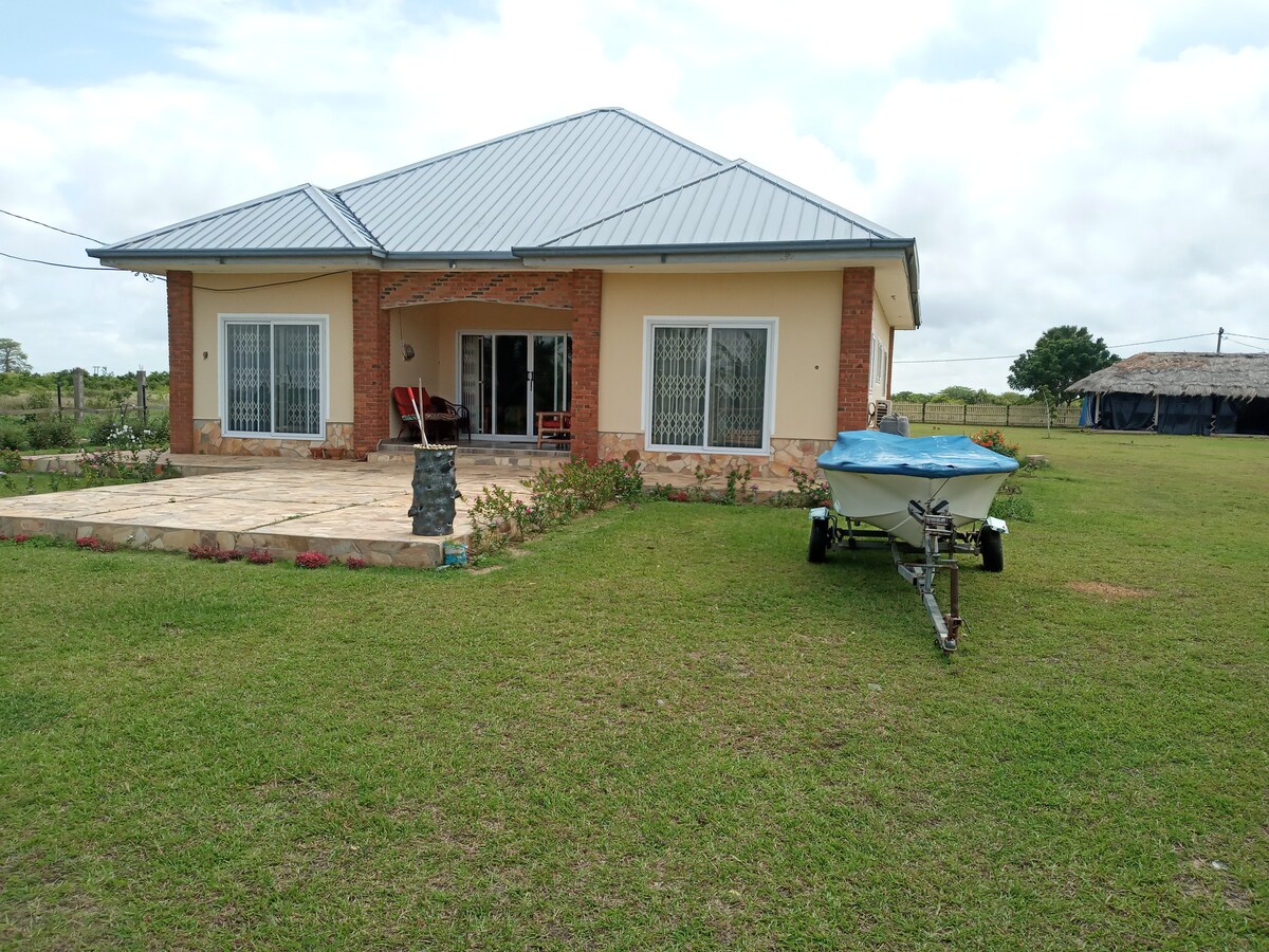 A single-story house with a modern design features a sloped metal roof and large glass doors opening to a spacious patio. A boat on a trailer is parked on well-maintained grass, with gardens and a distant structure visible beyond the property.