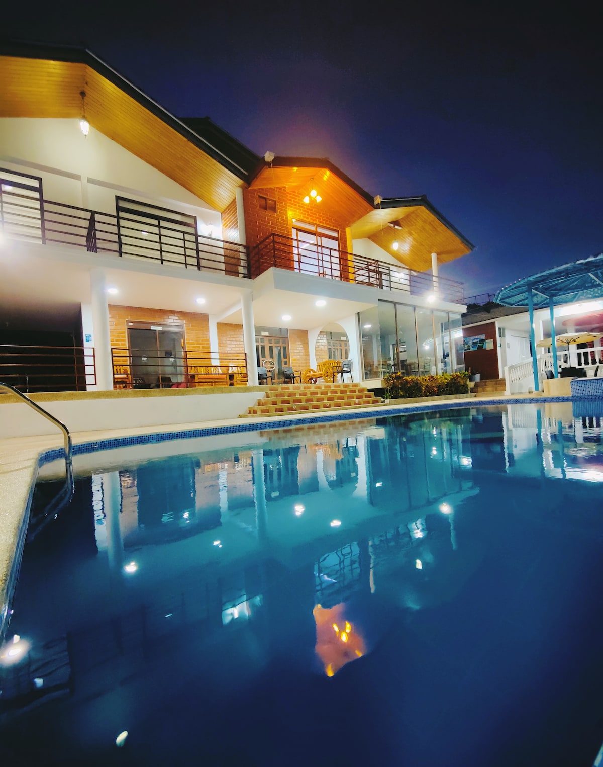 The exterior of the hostería is illuminated at night, reflecting warm lights off the surface of a tranquil pool. A two-story building features a combination of stone and wood accents, with multiple balconies and large windows showcasing inviting spaces.