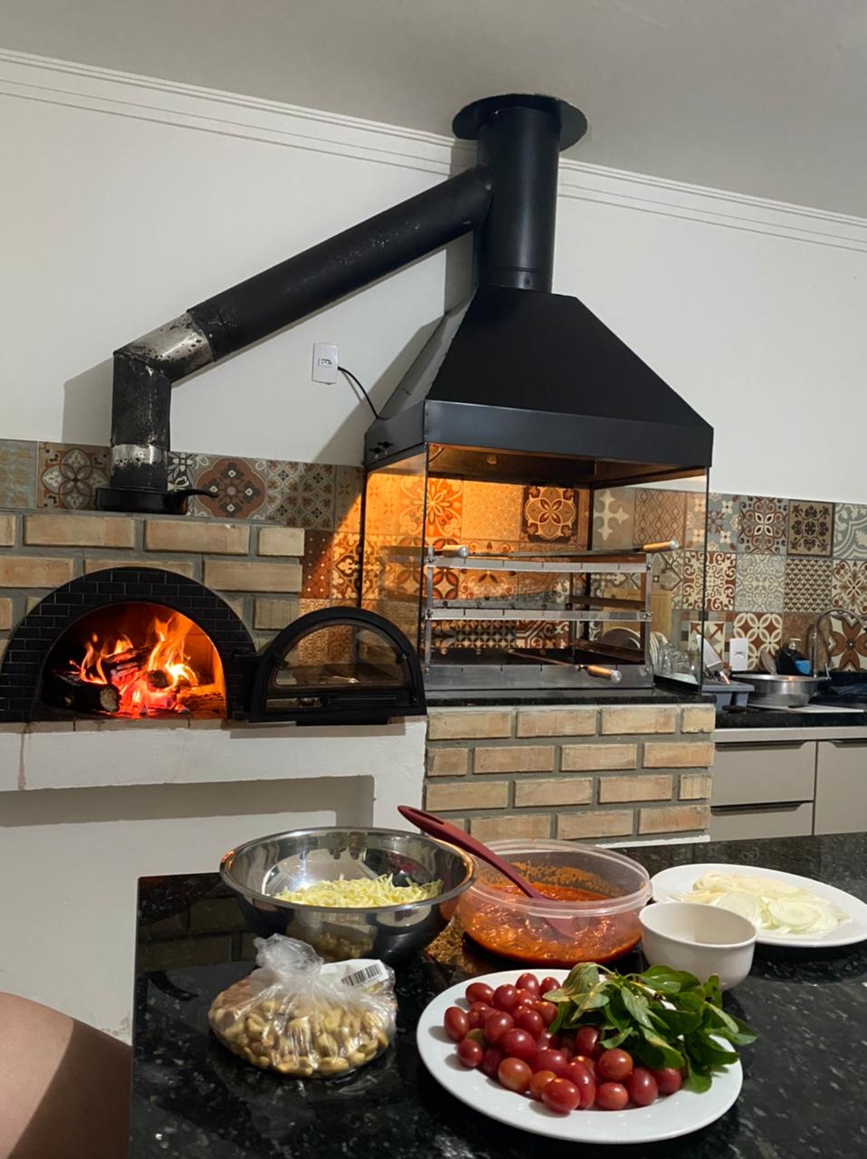 A gourmet kitchen area features a traditional wood-fired oven and an elegant chimney. Fresh ingredients, including tomatoes and herbs, are displayed on a dark countertop. A vibrant tiled backsplash adds texture and color to the space, enhancing the cooking environment.