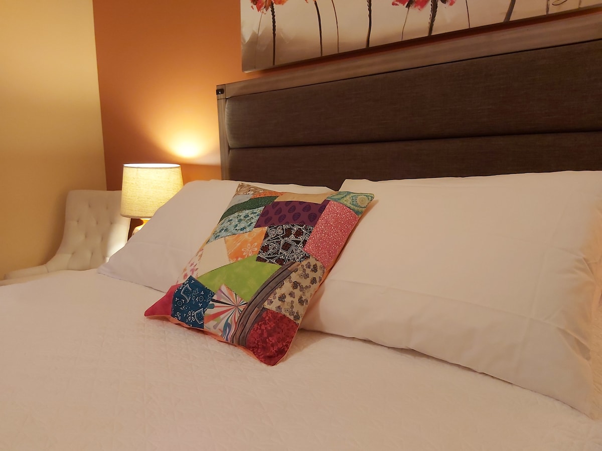 A well-appointed Queen size bed is presented with crisp white linens and decorative pillows. A patchwork pillow adds a splash of color. Soft light from a nearby lamp creates a warm ambiance, complementing the warm-toned walls adorned with floral artwork.