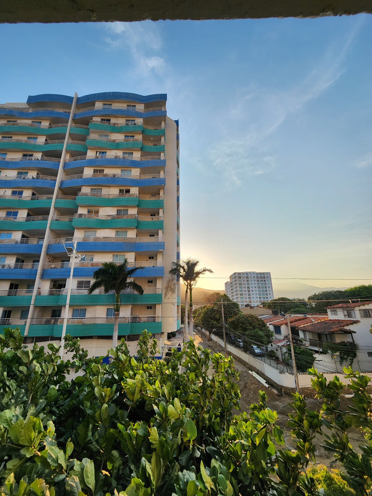 The exterior view showcases the tall residential building featuring multiple balconies with turquoise railings, illuminated by the soft glow of the setting sun. Lush green foliage frames the foreground, enhancing the sense of a serene environment in a vibrant location.
