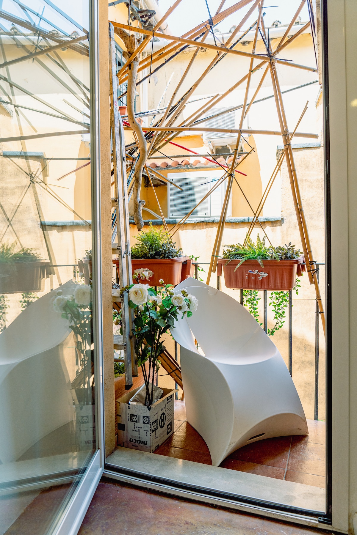 A small balcony is adorned with potted plants and features a modern, white chair. Natural light filters through the glass doors, illuminating the space. The balcony is enclosed by a unique arrangement of bamboo, adding an interesting visual element to the setting.
