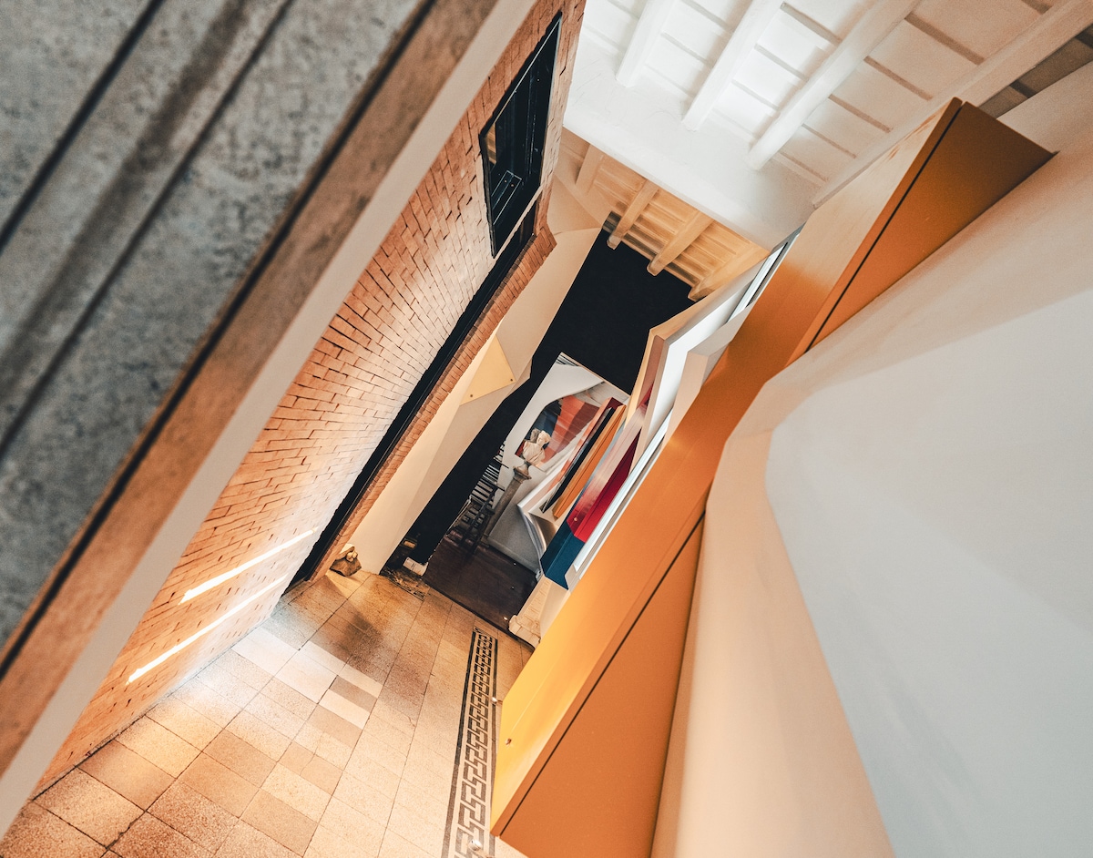 A hallway features a mix of textured walls, including exposed brick and smooth surfaces. Soft lighting highlights the architectural elements, and vibrant artwork peeks from a distance, creating a unique contrast within the space.