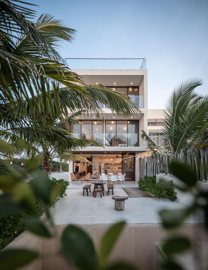 A modern three-story house is framed by lush palm trees, showcasing large glass windows and balconies. An outdoor terrace with seating is visible, creating a connection to the natural surroundings. The soft evening light enhances the inviting appearance of the property.