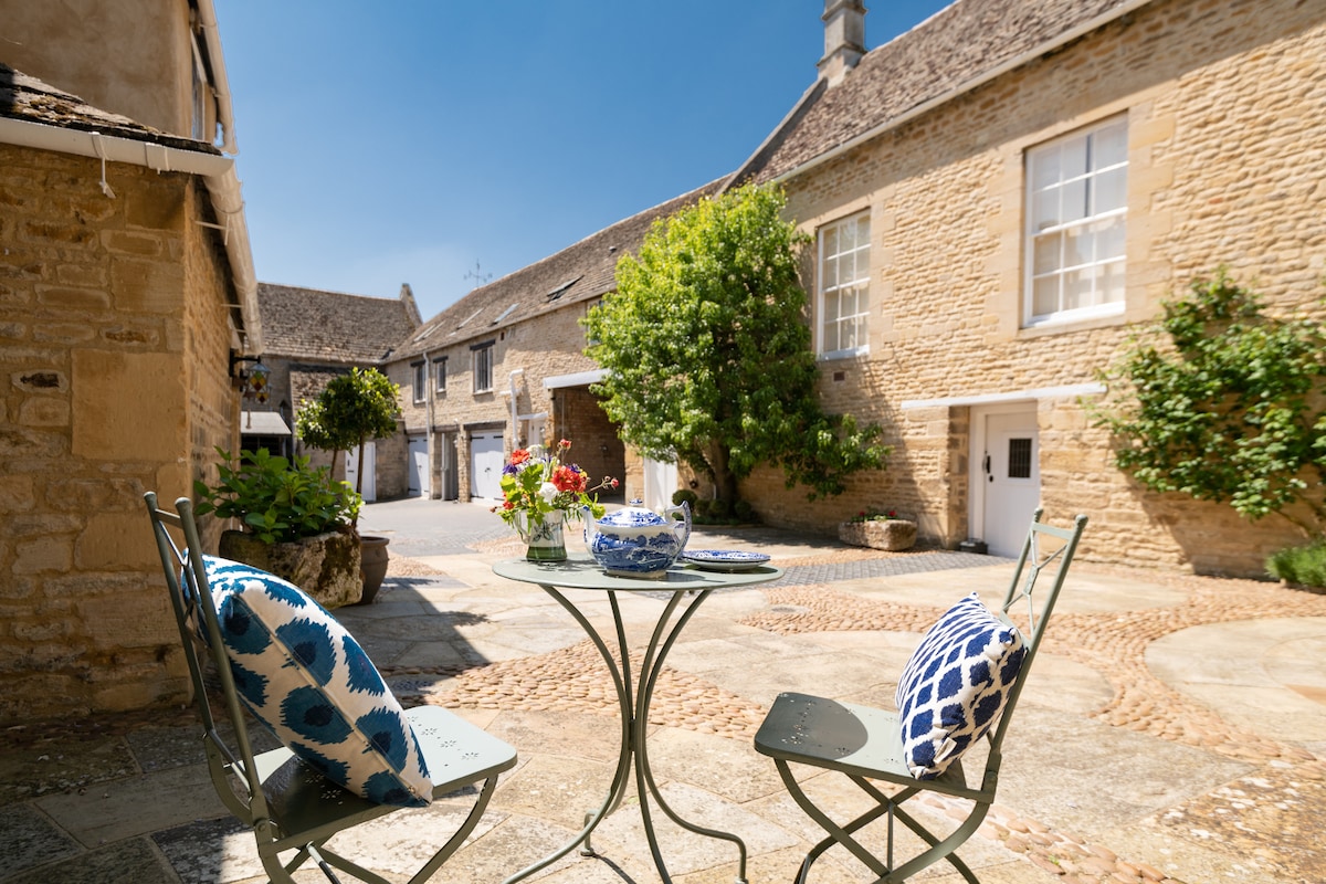 A charming courtyard features a small round table set with two chairs adorned with decorative cushions. Lush greenery provides a pleasant backdrop, while the textured stone buildings add character to the serene space. Sunlight creates a bright and inviting ambiance.