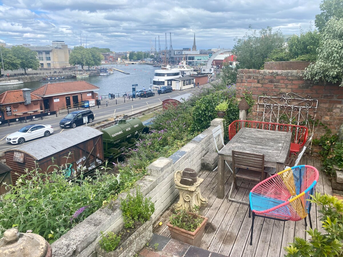 An outdoor seating area is depicted, featuring colorful chairs and a wooden table surrounded by greenery. The view overlooks the harbor, highlighting boats along the water and the city skyline under a cloudy sky. Decorative pots and plants add character to the space.