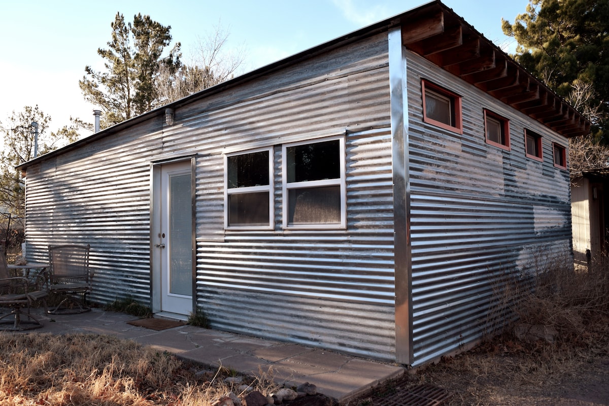 The exterior of the casita is showcased, featuring a unique corrugated metal façade with a simple wooden overhang. Light-colored double doors are positioned at the center, flanked by several windows. A small patio area with metal chairs is visible, surrounded by natural landscaping.