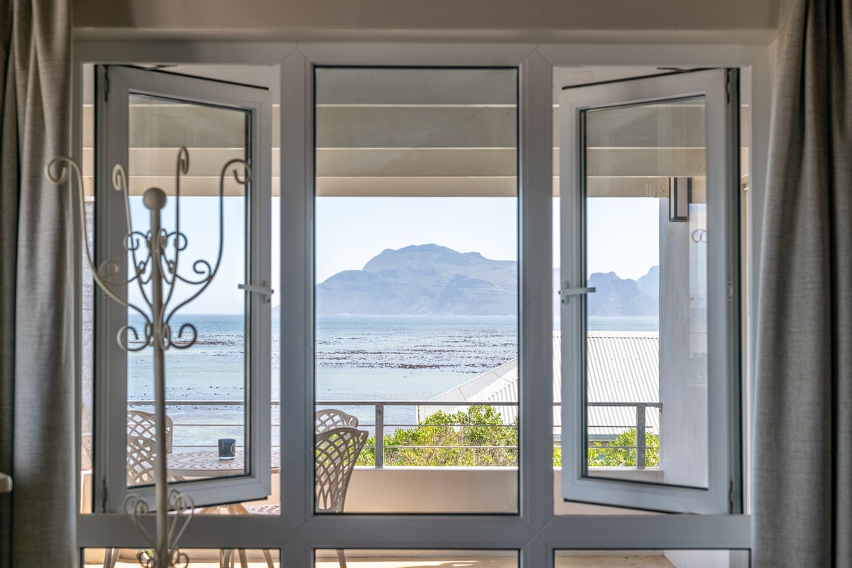 Large glass doors are opened wide, revealing an expansive view of the ocean and mountains. A small outdoor seating area is visible, featuring a round table and chairs. Light curtains frame the windows, enhancing the serene coastal atmosphere.