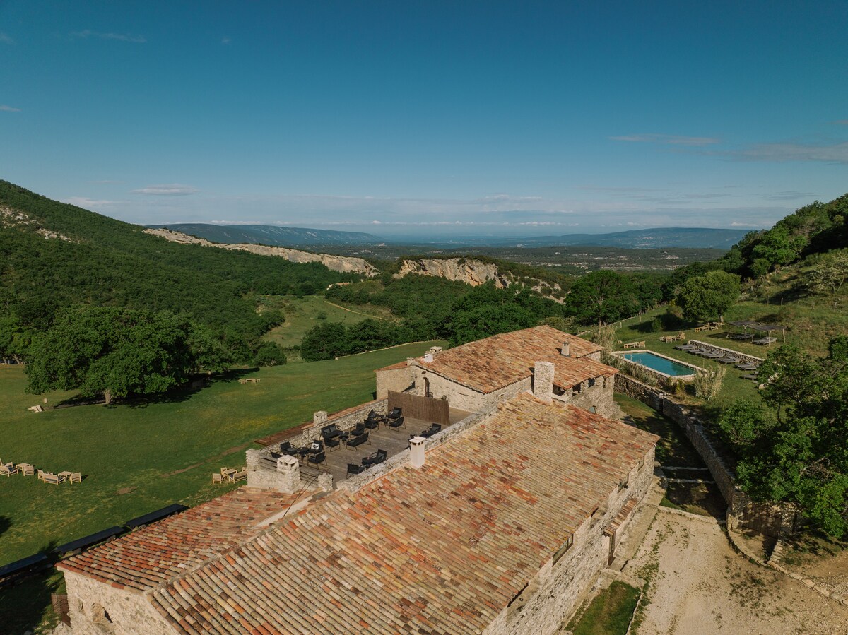 An expansive view of the property showcases the rustic stone architecture and tiled roof of the building. A spacious outdoor area includes a swimming pool surrounded by green fields and trees, with hills visible in the background under a clear blue sky.