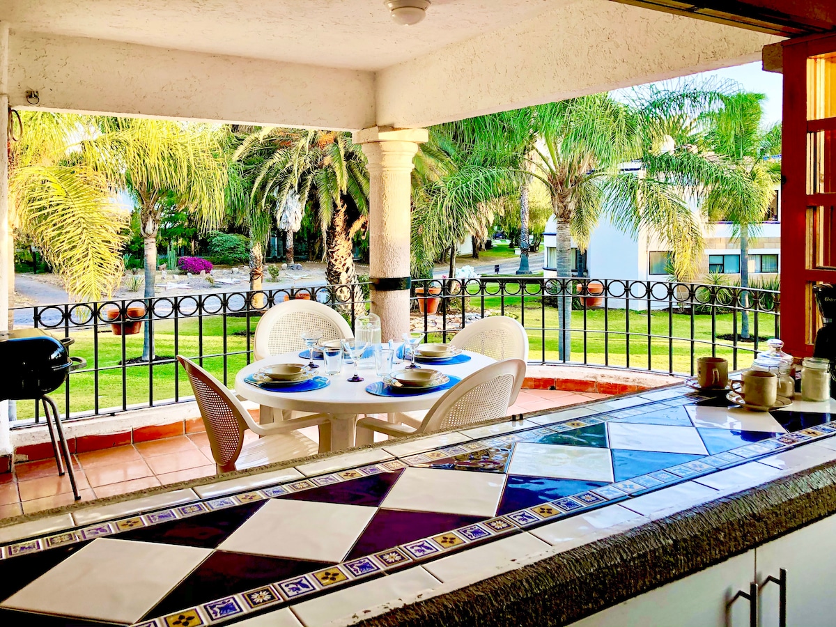 A bright terrace is visible through an open window, furnished with a round table and chairs for six. Lush palm trees and greenery line the view, while a barbecue grill is positioned at the edge of the space, adding to the inviting atmosphere.