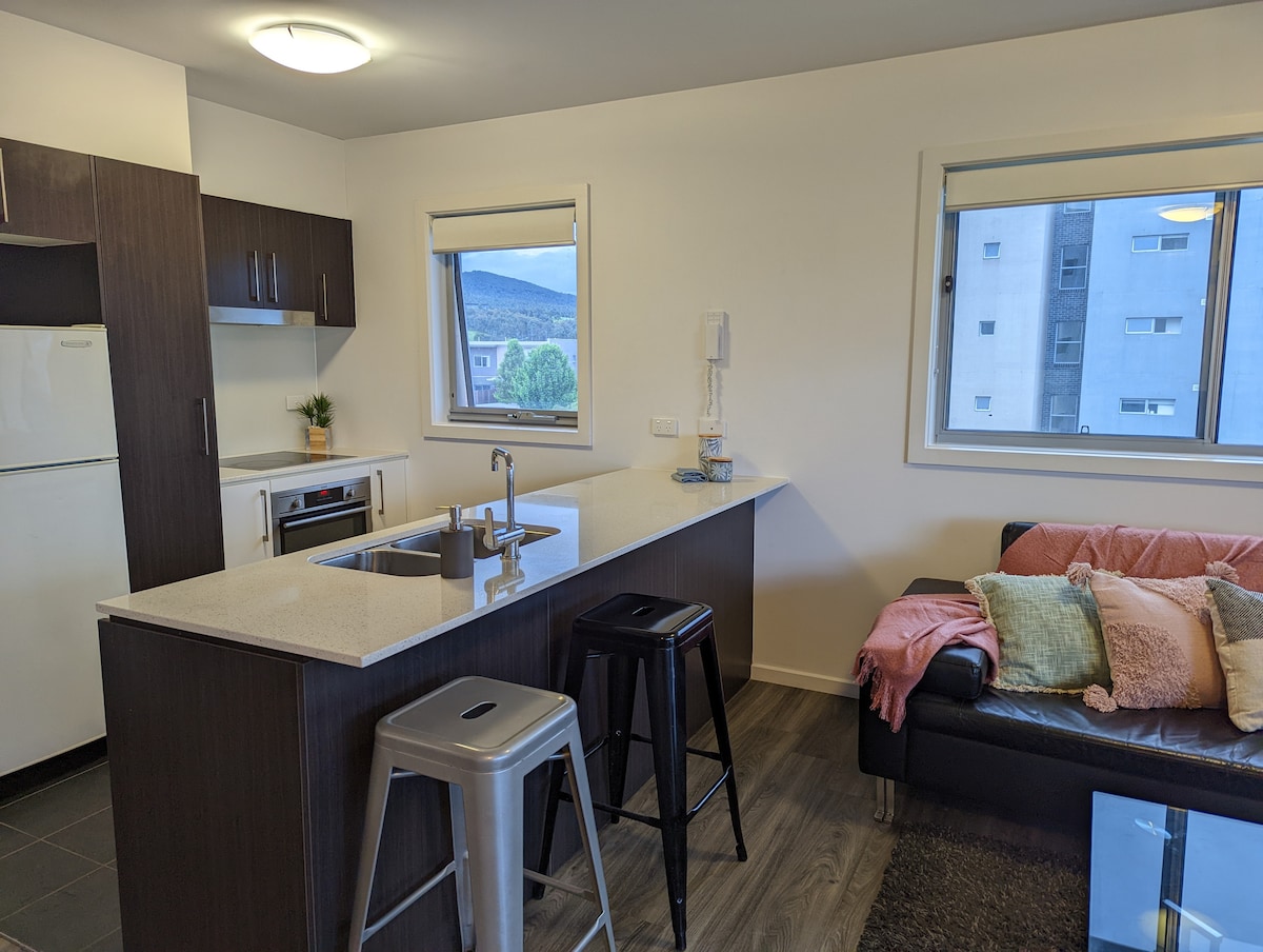 A modern kitchen area is featured with dark cabinetry and a light countertop. Two windows allow natural light, offering views of the surrounding landscape. A small dining island with bar stools is present, alongside a cozy seating arrangement with a colorful throw blanket.