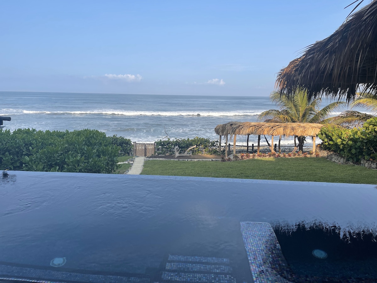 An infinity pool extends into the beachfront, providing sweeping views of the ocean. Gentle waves can be observed breaking against the shore. Lush greenery frames the area, with a thatched-roof structure visible in the distance alongside a sandy path.