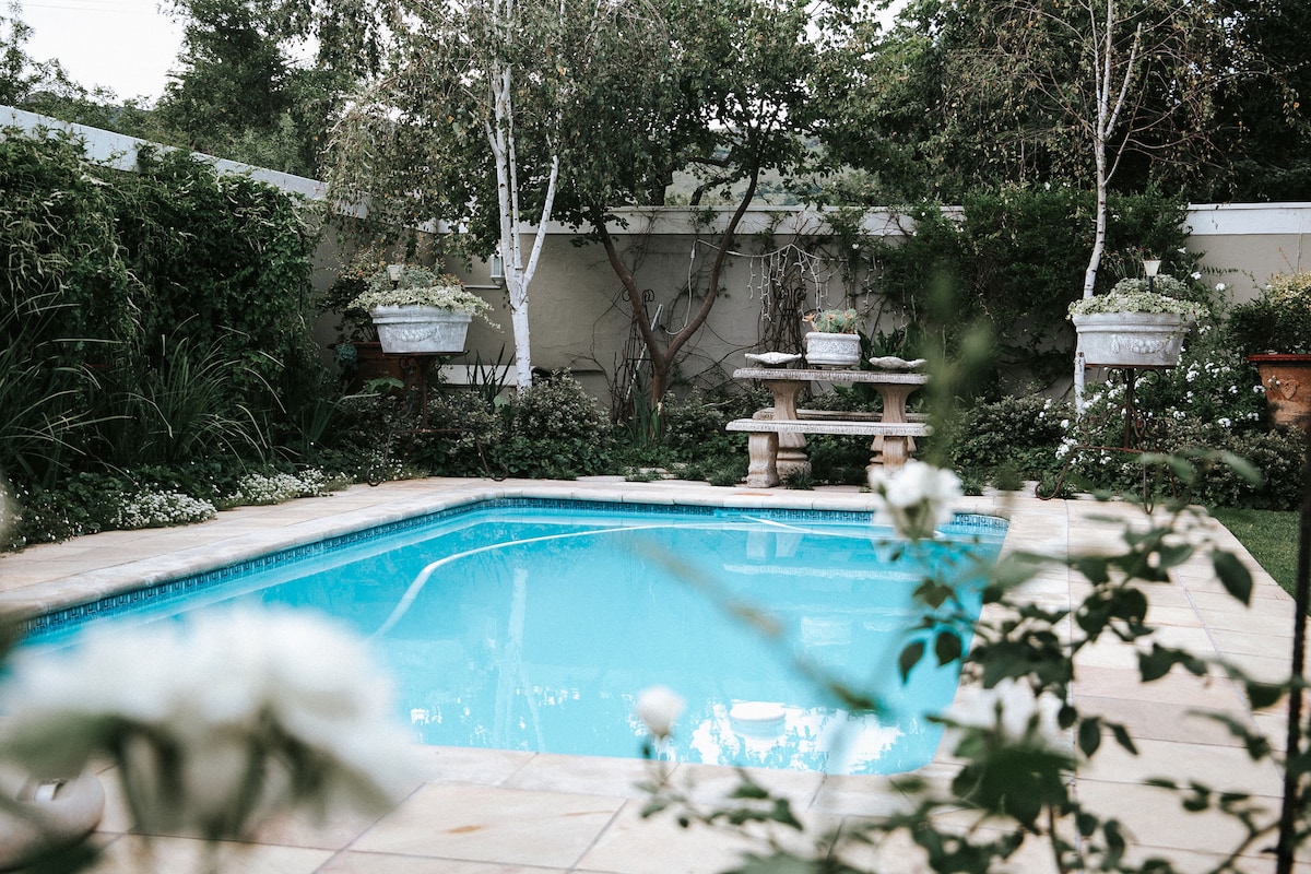 A serene swimming pool is surrounded by lush greenery and flowering plants. The clear blue water reflects the surrounding nature, while a stone table and benches are positioned nearby for relaxation.