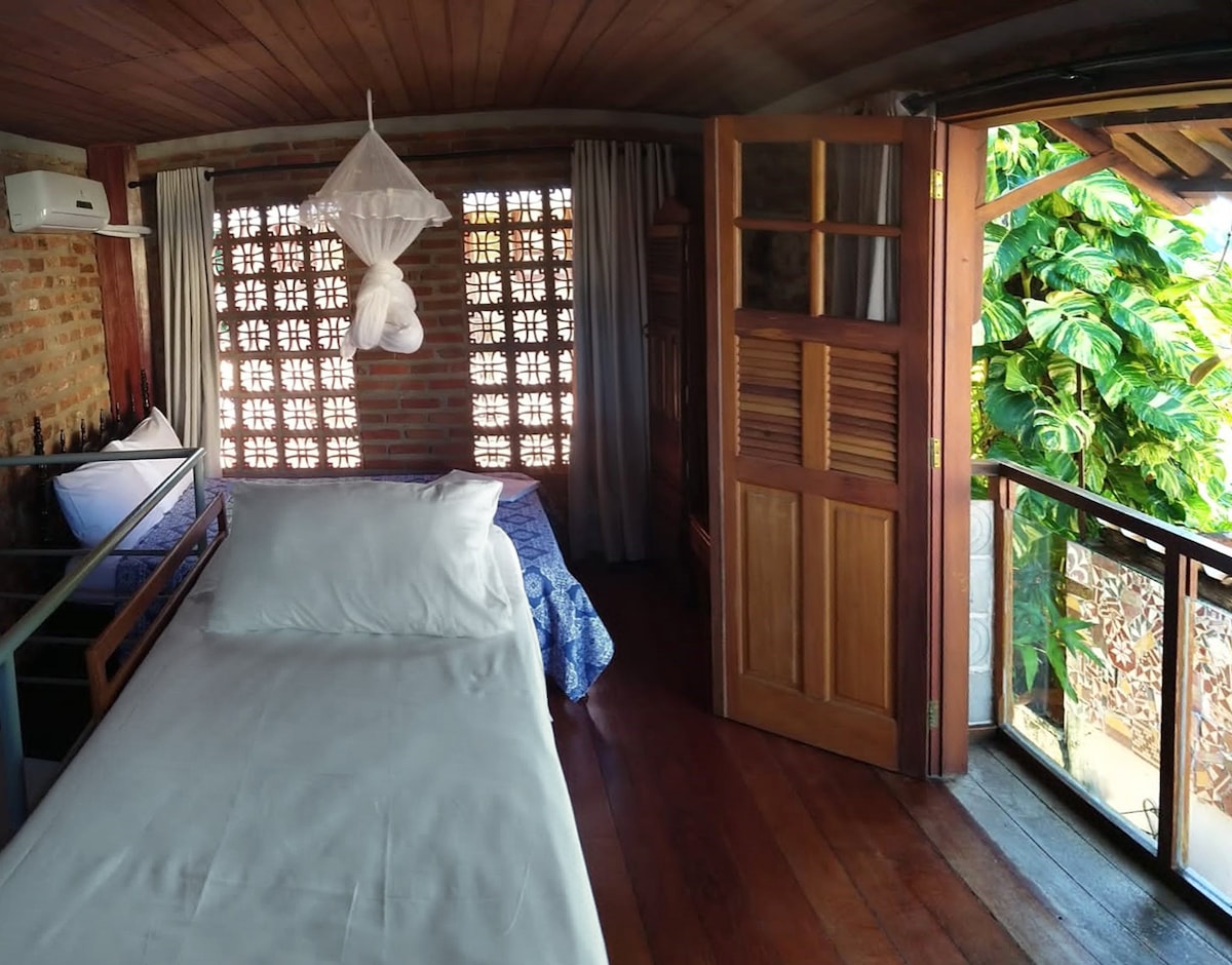 A bedroom features warm wooden flooring and rustic brick walls. A spacious bed is positioned beside a light blue patterned bedspread on an adjacent bed. Large windows allow natural light to fill the room, which opens to greenery outside.