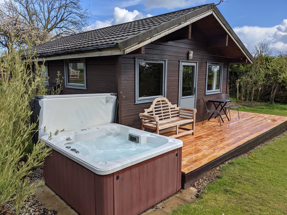 A wooden cabin features a welcoming hot tub on the deck, surrounded by greenery. Large windows offer views into the interior, while seating is provided on the porch with a wooden bench and a small table. The roof is slanted and overhanging.