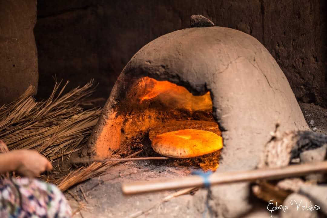 An adobe oven is shown, emitting a soft glow from the heat within. Traditional flatbread is placed inside, surrounded by dry brush and natural materials. The rustic texture of the oven suggests a connection to local culinary practices.