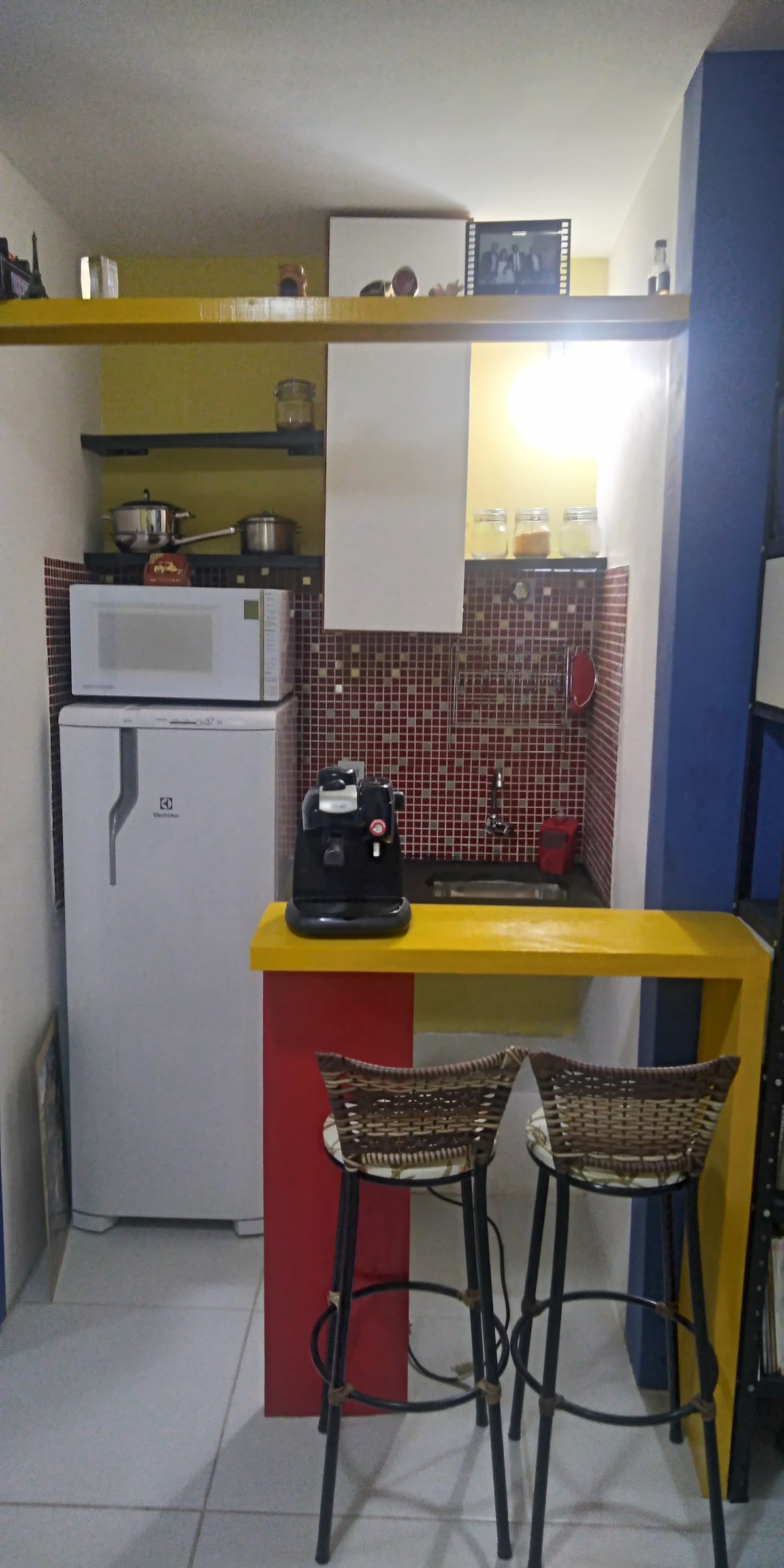 A compact kitchen area features a red and yellow color scheme, with modern appliances including a refrigerator and coffee maker. Bar stools provide seating at the small counter. Shelving above displays kitchenware, while the tiled backsplash adds a touch of color.