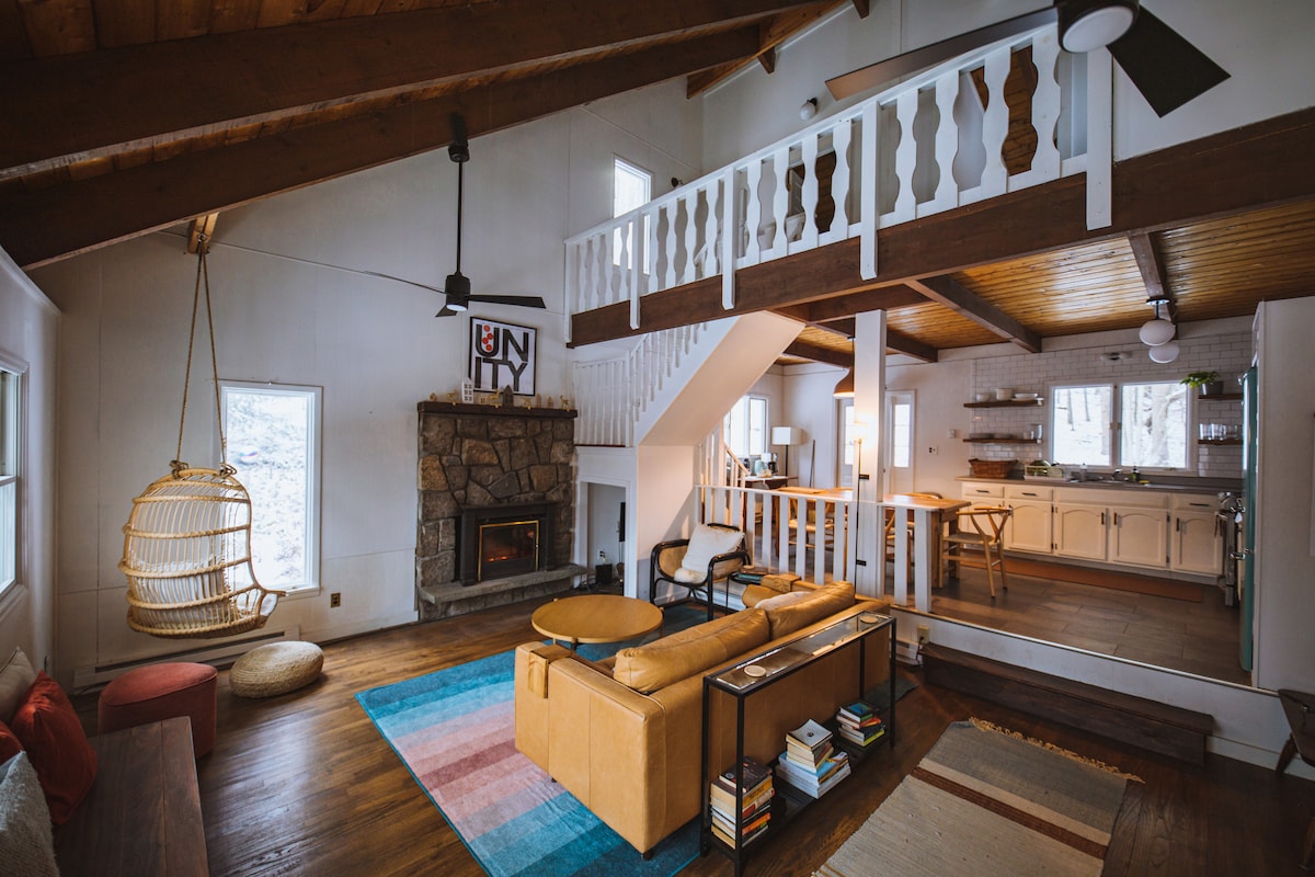 A spacious living area with vaulted ceilings features a stone fireplace and a hanging chair. Natural light enters from multiple windows, illuminating the wooden floors. A seating arrangement is centered around a coffee table, with a colorful area rug providing warmth.