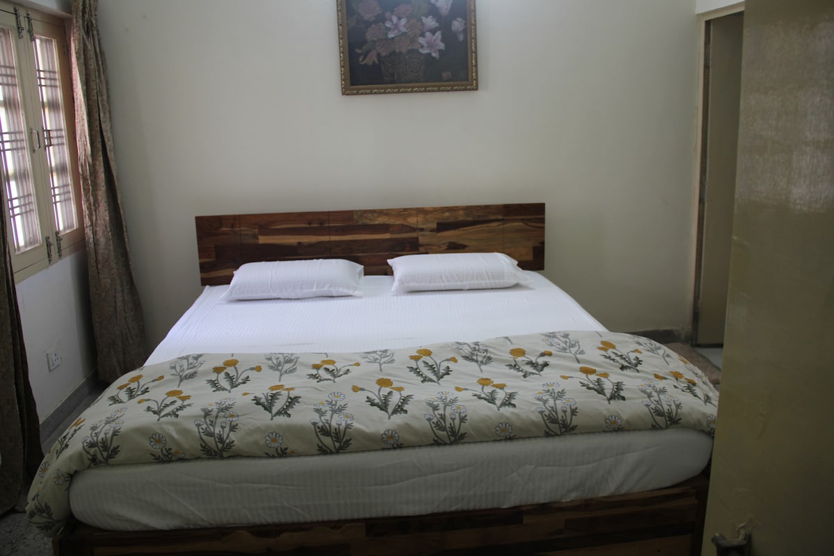 A serene bedroom features a spacious bed with crisp white linens and two neatly arranged pillows. A floral patterned throw blanket adds a touch of color. Natural light enters through a window adorned with light curtains, while a wall art piece enhances the overall ambiance.