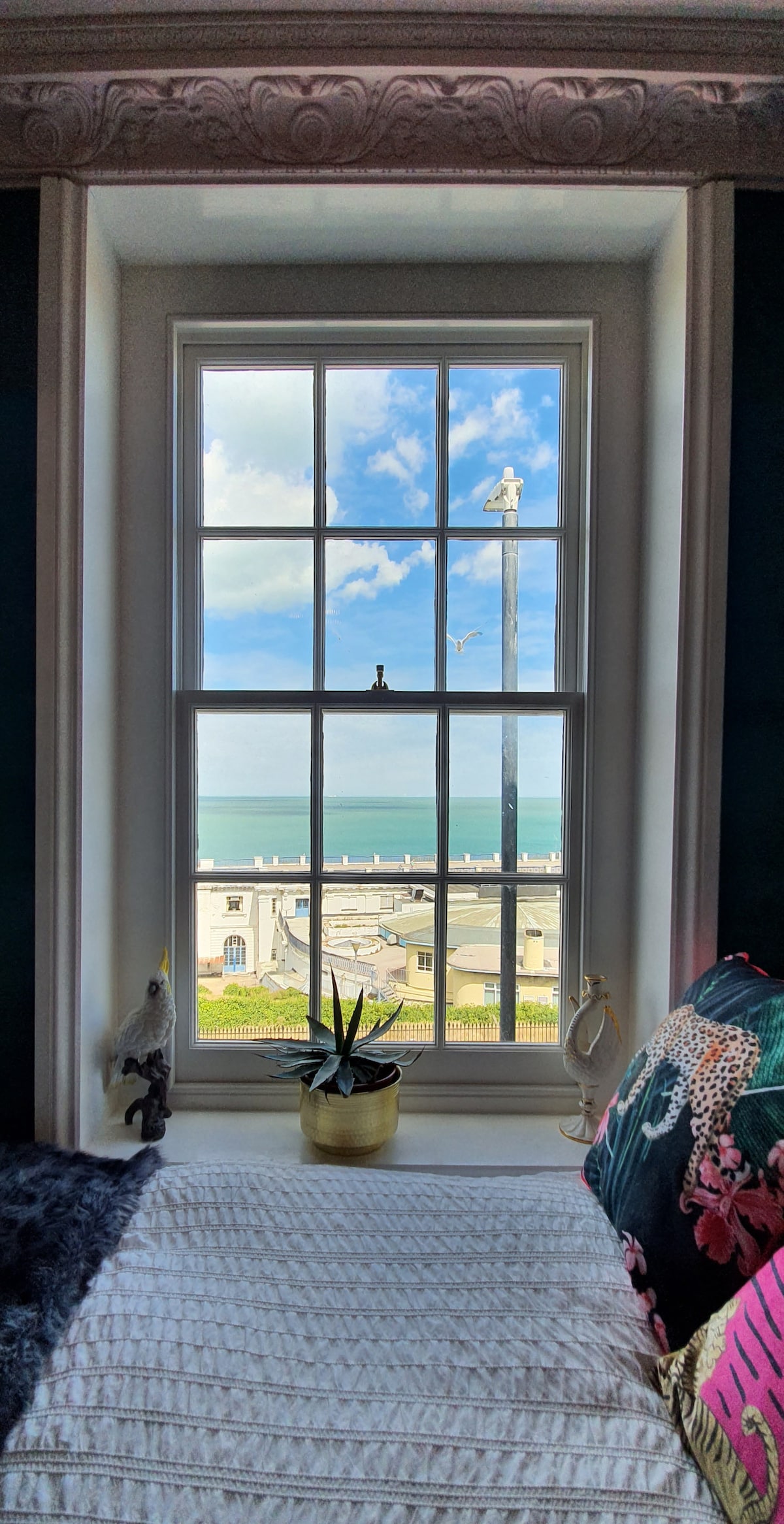 A window framed by ornate trim offers a view of the sea and a distant promenade. A potted plant sits on the windowsill, complementing the colorful cushions on the bed. Soft natural light fills the space, creating a serene ambiance.