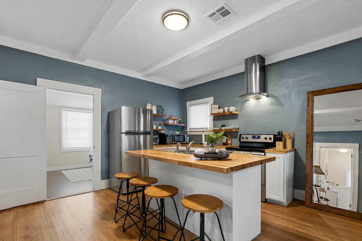 A modern kitchen features a central wooden island with three stools, offering a casual dining spot. Stainless steel appliances, including a refrigerator and stove, are visible against a blue wall. Natural light filters in through nearby windows, complementing the clean and functional layout.
