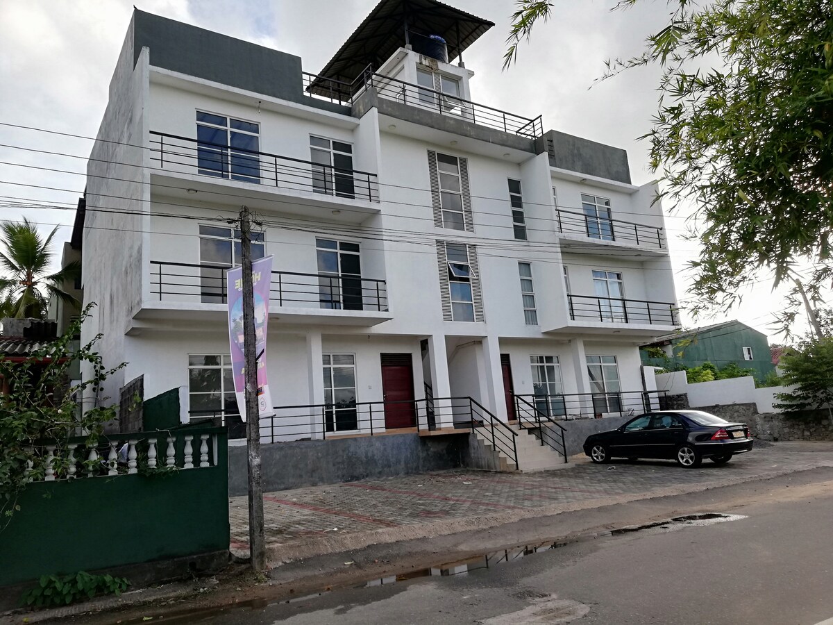 The modern apartment building features three levels with a clean, white exterior. Large windows provide ample natural light. A sloped roof extends over the top, and a paved pathway leads to the main entrance, accessible via a set of steps.