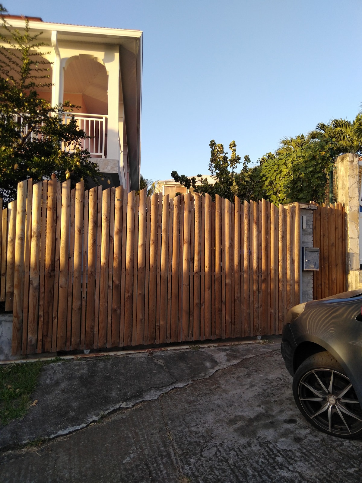 A wooden gate, crafted from vertical slats, stands closed at the entrance of a property. The surrounding area is landscaped with greenery, and a car is parked nearby on a concrete driveway. The sky is clear and blue in the background.
