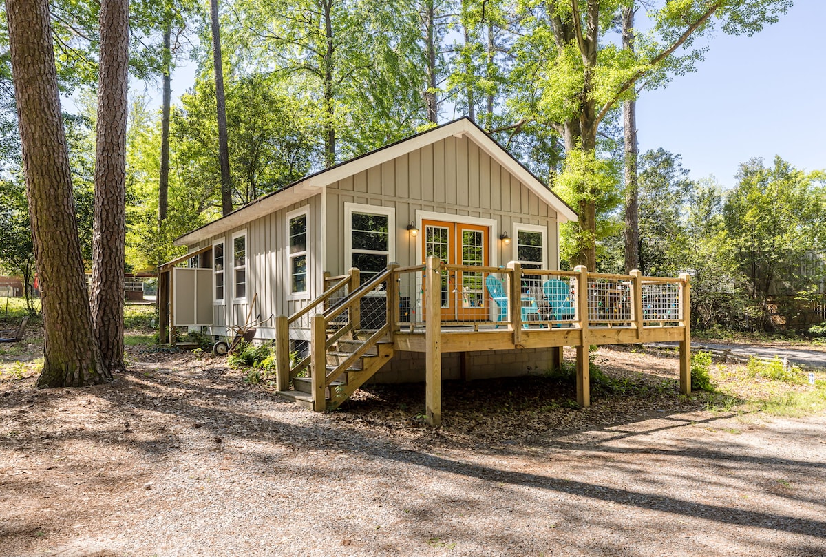 The cottage is surrounded by trees, featuring a wooden deck with seating. Glass doors provide access to the interior, while the exterior showcases a blend of light and dark siding. A gravel pathway leads up to the entrance, enhancing the natural setting.
