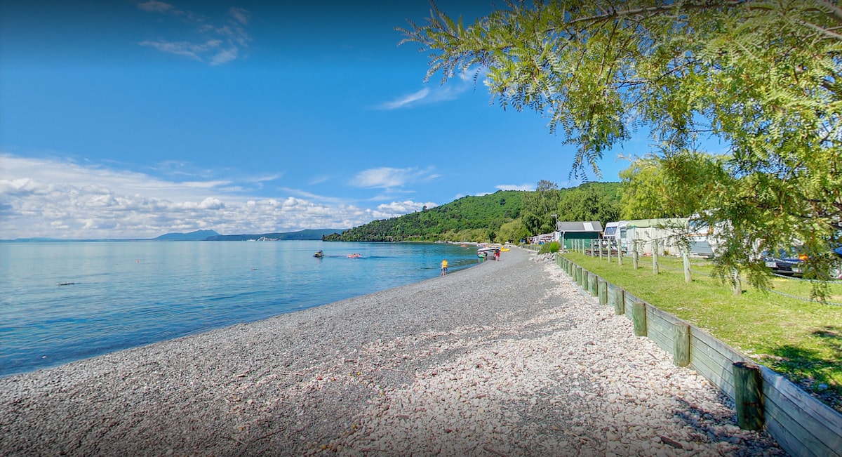 A scenic shoreline is showcased, featuring smooth pebbles leading to clear waters. Lush greenery and hills are visible in the background, while boats can be seen along the water's edge. The atmosphere appears tranquil, inviting leisurely walks along the beach.