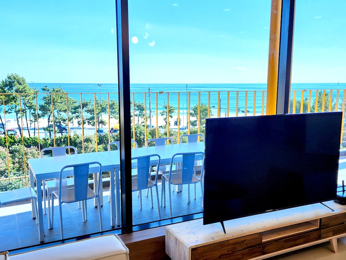 A spacious living area offers a clear ocean view through large glass windows. A 65-inch TV is positioned in front of a modern wooden entertainment unit. A dining table for ten is set on the balcony, inviting guests to enjoy meals with scenic vistas.