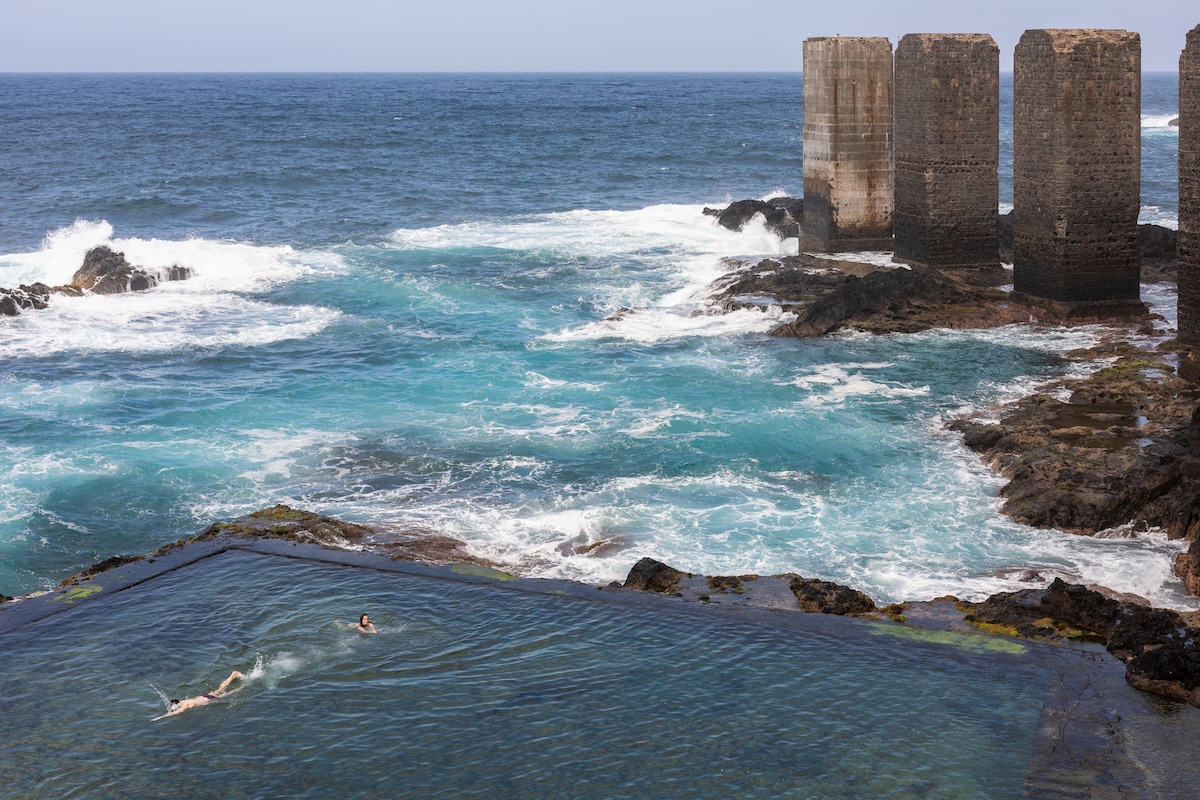 A serene ocean scene features two individuals swimming in a natural pool framed by rocky shores, with waves crashing against the coastline. The blue waters reflect the sky, while stone pillars of an old structure stand prominently in the background amidst the vibrant sea.