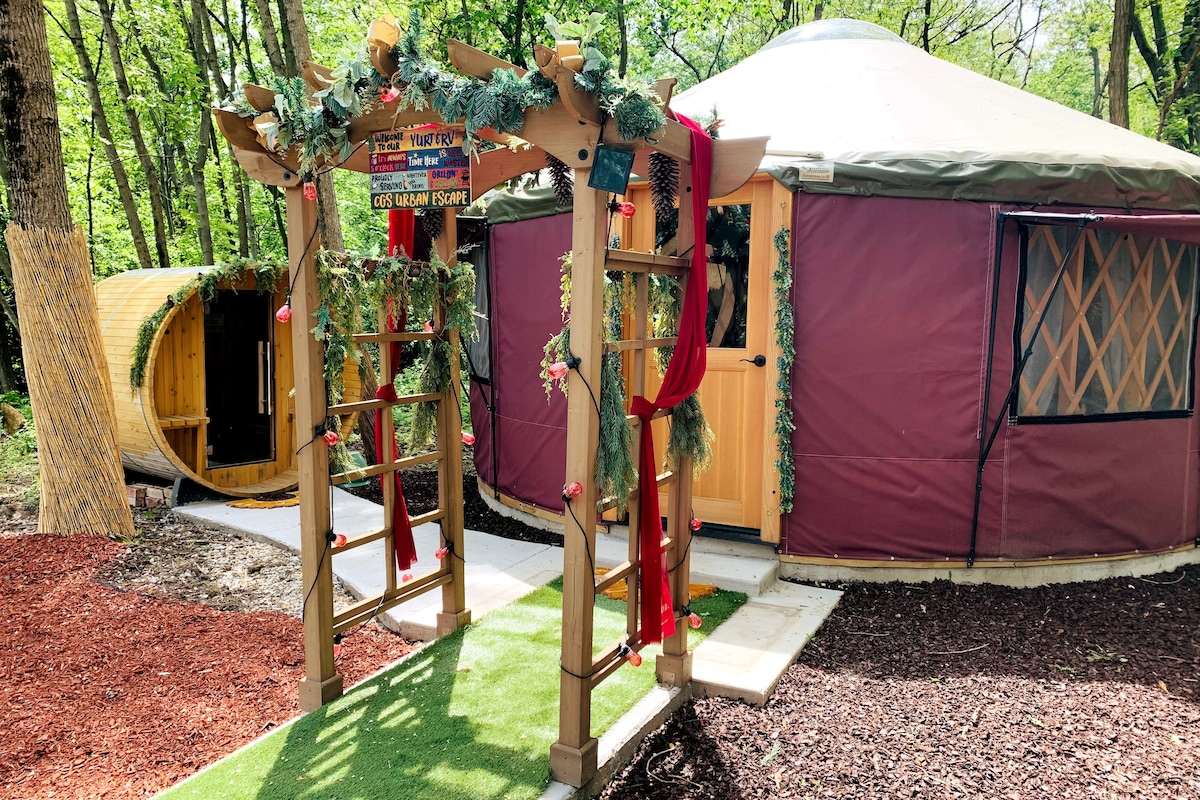 A natural pathway leads to a charming safari yurt, adorned with greenery and decorative elements. A wooden archway introduces the entrance, while a nearby barrel sauna complements the serene outdoor setting, surrounded by trees and mulch landscaping.
